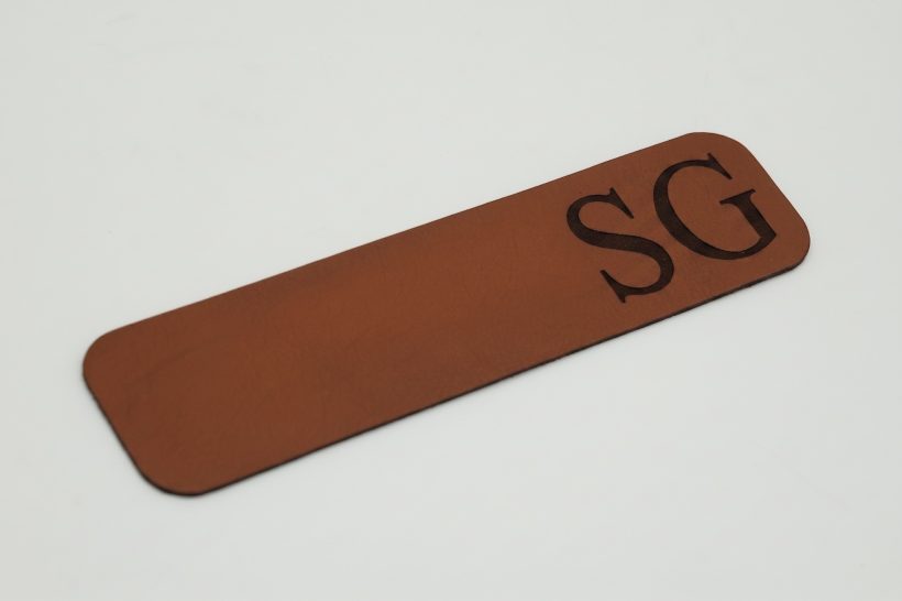 Personalisiertes Lesezeichen aus Echtleder mit Lasergravur Braunes Echtleder-Lesezeichen mit Lasergravur der Initialen SG und abgerundeten Ecken.